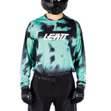 A long-sleeve motocross jersey with a teal and black tie-dye pattern, featuring the LEATT logo on the chest, modeled with black pants, gloves, and a black helmet against a white background.